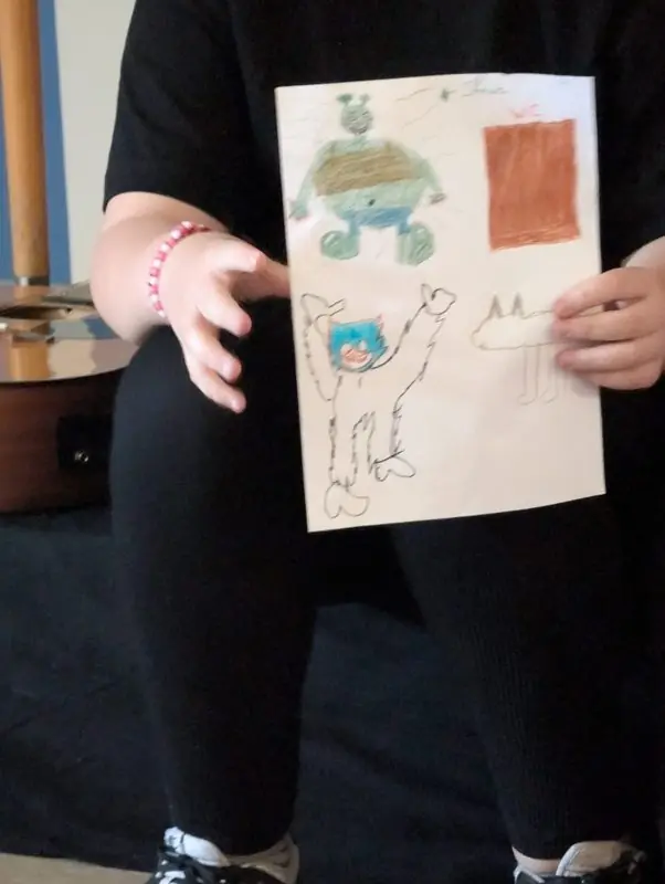 Un enfant montre le dessin de son conte : des personnages colorés et une maison.